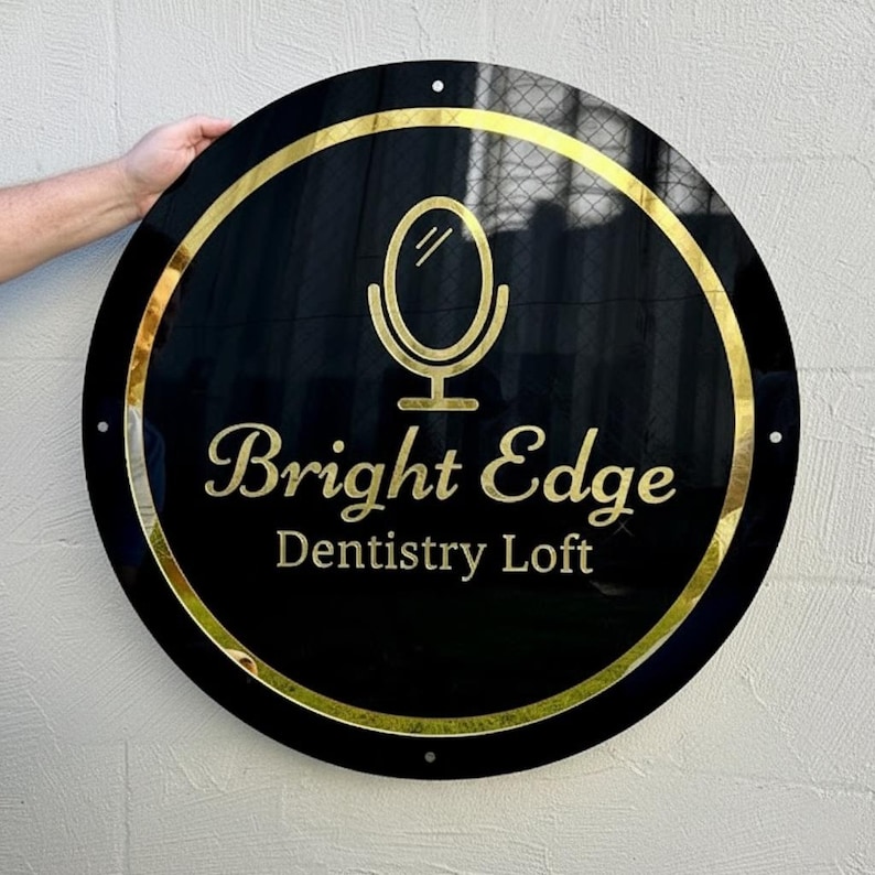 May include: A round, black sign with gold trim and lettering. The sign reads "Bright Edge Dentistry Loft" in gold script. A gold mirror icon is above the text. The sign is held by a person's hand.