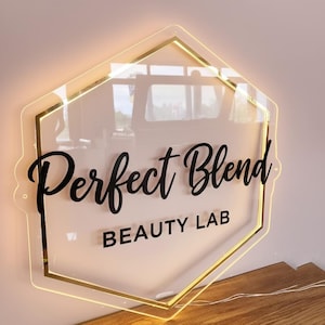 May include: A clear acrylic sign with the words "Perfect Blend BEAUTY LAB" in black script and block letters. The sign is in a gold-lined, hexagon shape with a warm, glowing light around the edges. The sign is mounted on a light pink wall.