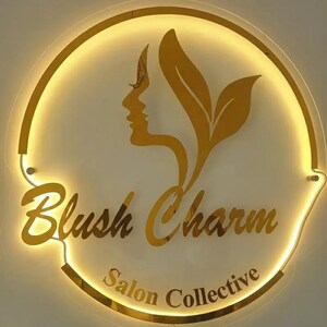 May include: A gold-colored, illuminated sign with the words "Blush Charm Salon Collective." The sign features a silhouette of a woman's face and stylized leaves. The sign is circular with a gold border.