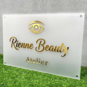 May include: A rectangular white sign with gold lettering and a gold eye logo. The sign reads "Rienne Beauty Atelier". The sign is mounted on a green surface.
