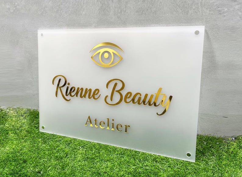 May include: A rectangular white sign with gold lettering and a gold eye logo. The sign reads "Rienne Beauty Atelier". The sign is mounted on a green surface.
