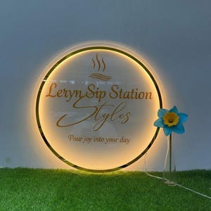 Puede incluir: Un letrero redondo iluminado con el texto "Leryn Sip Station Styles" en letras doradas. Una flor azul y amarilla en un jarr&oacute;n de cristal est&aacute; a la derecha del letrero. El letrero tiene un borde dorado y est&aacute; sobre un lecho de hierba verde.
