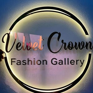 May include: A circular sign with the words "Velvet Crown Fashion Gallery" in a stylish font. The sign has a black border with a warm, glowing light around the edge. The words "Velvet" and "Crown" are in a rose gold color.