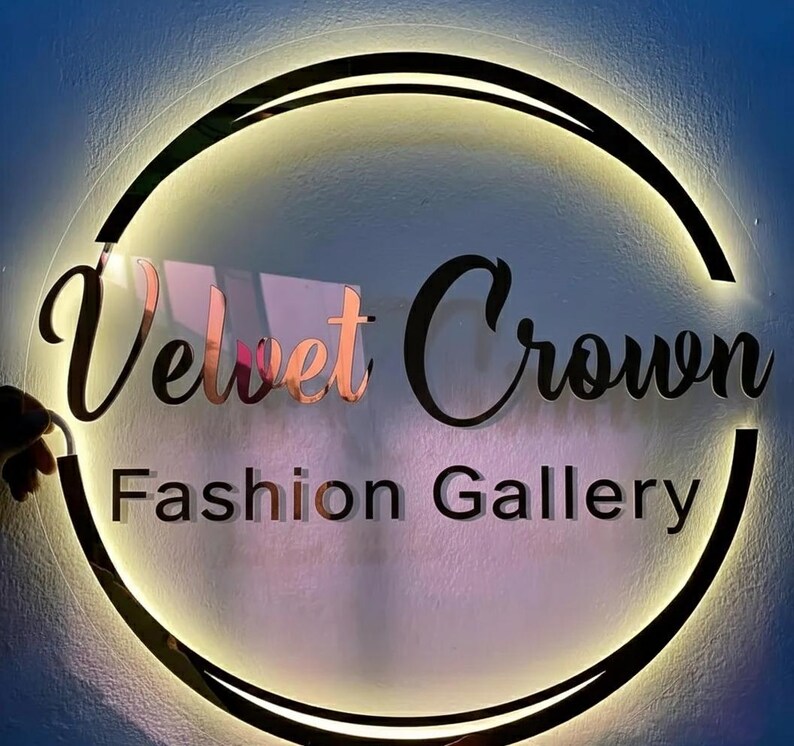 May include: A circular sign with the words "Velvet Crown Fashion Gallery" in a stylish font. The sign has a black border with a warm, glowing light around the edge. The words "Velvet" and "Crown" are in a rose gold color.