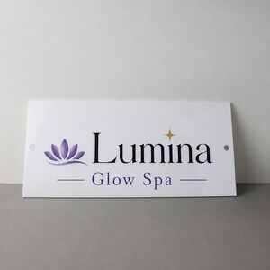 May include: White rectangular sign with the text "Lumina Glow Spa" in black and purple. The sign features a purple lotus flower graphic and a gold star. Two holes are visible on the sides.