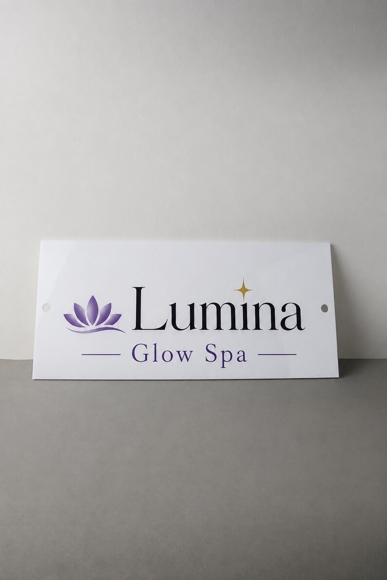 May include: White rectangular sign with the text "Lumina Glow Spa" in black and purple. The sign features a purple lotus flower graphic and a gold star. Two holes are visible on the sides.