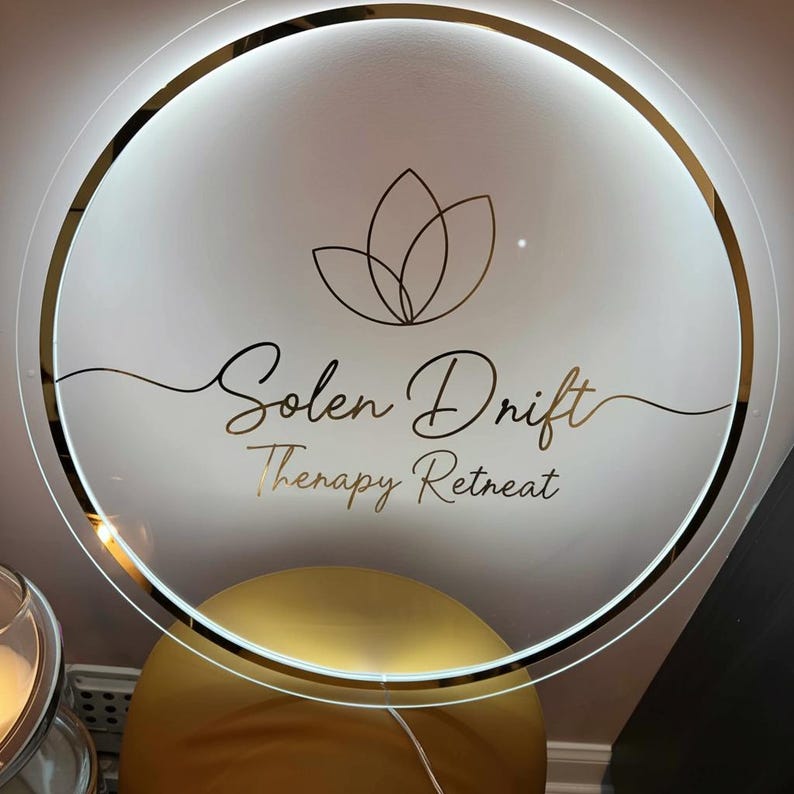 K&ouml;nnte beinhalten: Ein rundes, beleuchtetes Schild mit goldenem Rand und dem Schriftzug "Solen Drift Therapy Retreat" in eleganter Schrift. Das Schild zeigt oben ein goldenes Lotusblumen-Design. Der Hintergrund ist eine weiche, neutrale Farbe.