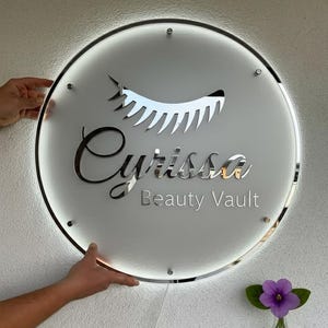 Puede incluir: Un letrero redondo e iluminado con el texto "Cyrissa Beauty Vault" en letras plateadas. El letrero presenta un dise&ntilde;o de pesta&ntilde;as estilizado y est&aacute; sostenido por manos. Un peque&ntilde;o jarr&oacute;n con una flor morada est&aacute; en la esquina inferior derecha.