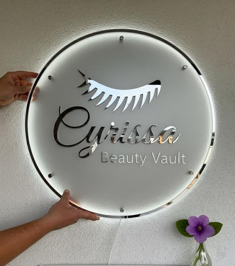 Puede incluir: Un letrero redondo e iluminado con el texto "Cyrissa Beauty Vault" en letras plateadas. El letrero presenta un dise&ntilde;o de pesta&ntilde;as estilizado y est&aacute; sostenido por manos. Un peque&ntilde;o jarr&oacute;n con una flor morada est&aacute; en la esquina inferior derecha.