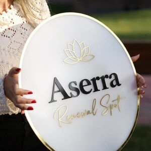 May include: A round white sign with gold trim and the text "Aserra Renewal Spot" in black and gold. A gold lotus flower design is at the top. The sign is held by a person.