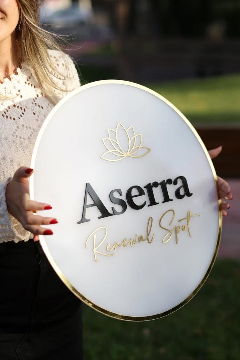 May include: A round white sign with gold trim and the text "Aserra Renewal Spot" in black and gold. A gold lotus flower design is at the top. The sign is held by a person.