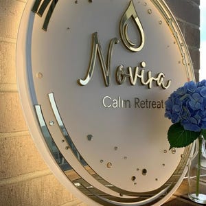 Puede incluir: Un letrero redondo e iluminado con el nombre "Novira" en letras doradas y las palabras "Calm Retreat" debajo. El letrero tiene un borde dorado y un dise&ntilde;o de gota de agua. A la derecha se ve un jarr&oacute;n con hortensias azules.