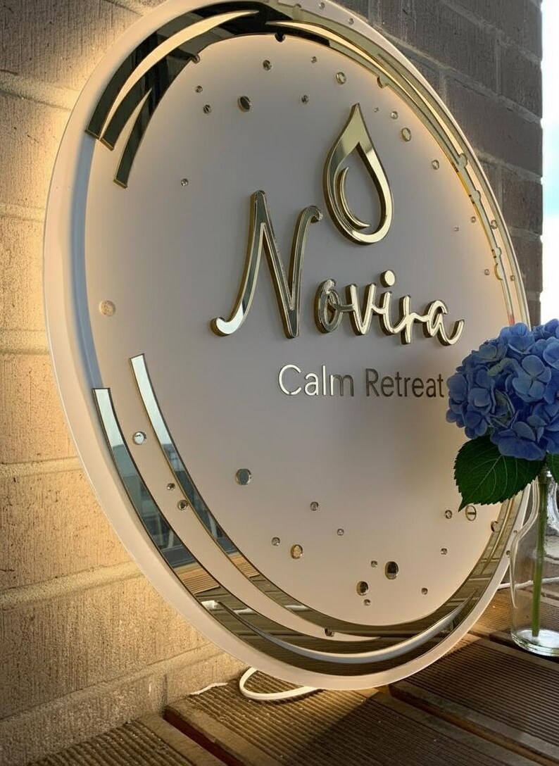 Puede incluir: Un letrero redondo e iluminado con el nombre "Novira" en letras doradas y las palabras "Calm Retreat" debajo. El letrero tiene un borde dorado y un dise&ntilde;o de gota de agua. A la derecha se ve un jarr&oacute;n con hortensias azules.