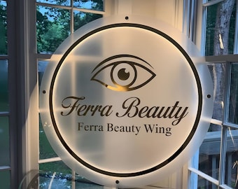 May include: A round, illuminated sign with the text "Ferra Beauty" and "Ferra Beauty Wing" in elegant script. The sign features a gold-colored eye graphic and a black border. The sign is mounted on a window.