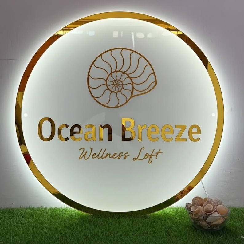 Pu&ograve; includere: Un cartello rotondo con bordo dorato e le parole "Ocean Breeze Wellness Loft" in lettere dorate. Un disegno di conchiglia dorata &egrave; sopra il testo. Il cartello &egrave; illuminato da una luce bianca. Una piccola ciotola di vetro con conchiglie &egrave; sotto.