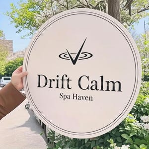 May include: A round, white sign with black text that reads "Drift Calm Spa Haven." The sign has a black border and a logo above the text. The logo is a checkmark inside a stylized water ripple design. The sign is held outdoors.