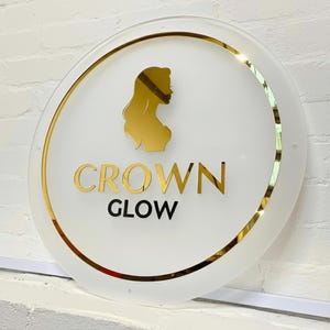 Pu&ograve; includere: Un cartello rotondo bianco con bordo dorato e una silhouette dorata del profilo di una donna. Le parole "CROWN GLOW" sono in lettere dorate. Il cartello &egrave; montato su un muro di mattoni bianchi.