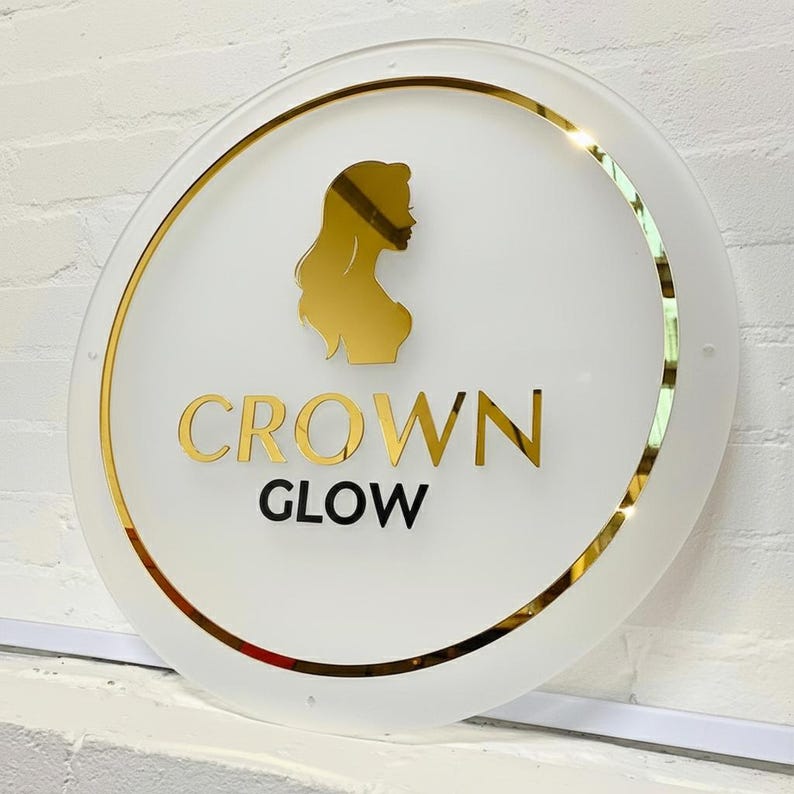 Pu&ograve; includere: Un cartello rotondo bianco con bordo dorato e una silhouette dorata del profilo di una donna. Le parole "CROWN GLOW" sono in lettere dorate. Il cartello &egrave; montato su un muro di mattoni bianchi.