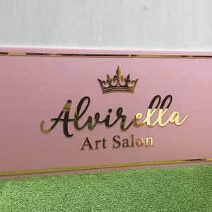 May include: A pink rectangular sign with gold trim and lettering. The sign reads "Alvirella Art Salon" in gold script and block letters, topped with a gold crown. The sign is displayed on a green surface.