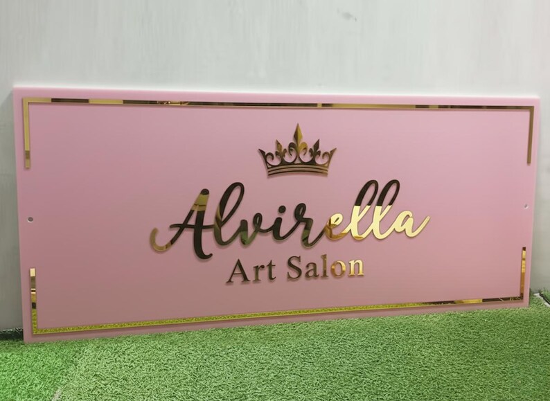 May include: A pink rectangular sign with gold trim and lettering. The sign reads "Alvirella Art Salon" in gold script and block letters, topped with a gold crown. The sign is displayed on a green surface.