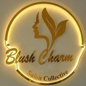 May include: A gold-coloured, illuminated sign with the words "Blush Charm Salon Collective." The sign features a silhouette of a woman's face and stylized leaves. The sign is circular with a gold border.