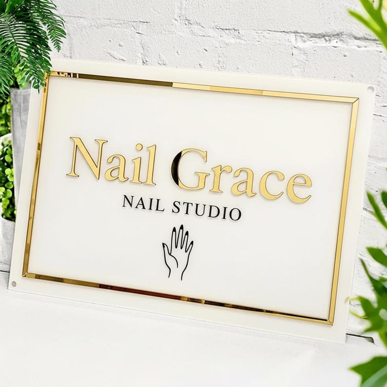 May include: A rectangular sign with a white background and gold trim. The words "Nail Grace" are in gold lettering, with "NAIL STUDIO" below in black. A black hand graphic is at the bottom.