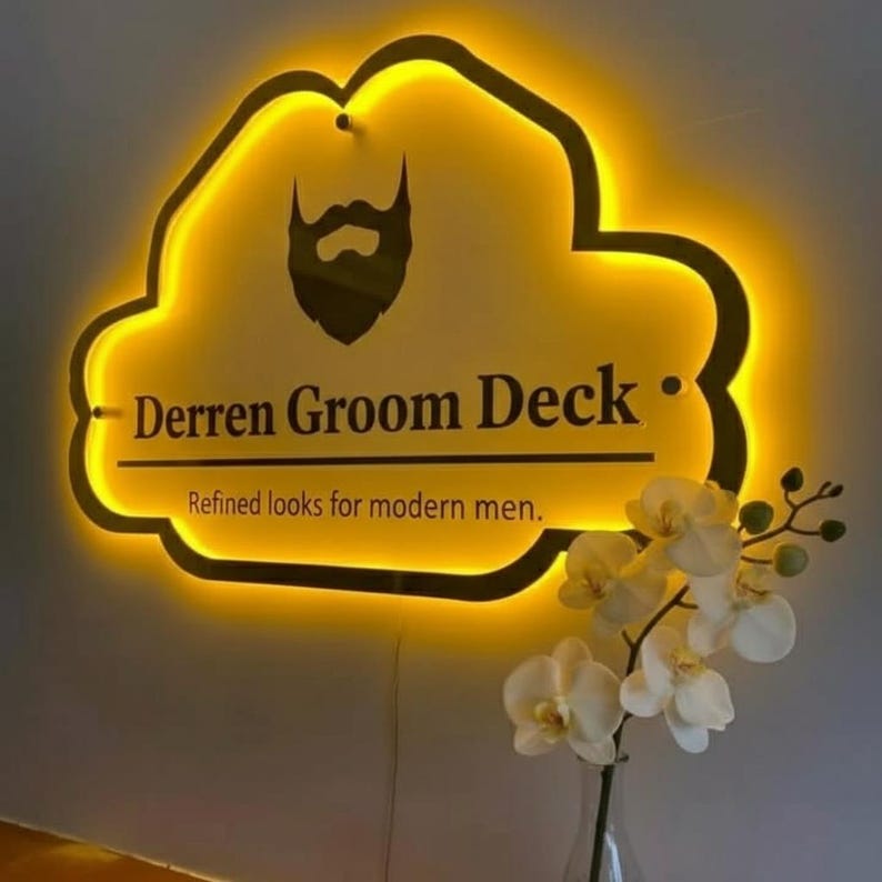 Puede incluir: Letrero luminoso con forma de nube con el texto "Derren Groom Deck" y un gr&aacute;fico de barba. El letrero tiene un brillo amarillo. El texto "Refined looks for modern men" est&aacute; debajo. Orqu&iacute;deas blancas en un jarr&oacute;n.