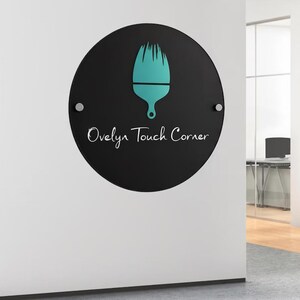 May include: A round, black sign with the text "Ovelyn Touch Corner" in white script. A teal-colored paintbrush icon is centered above the text. The sign is mounted on a white wall with two silver fasteners.
