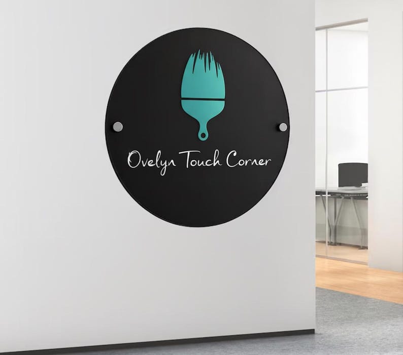 May include: A round, black sign with the text "Ovelyn Touch Corner" in white script. A teal-colored paintbrush icon is centered above the text. The sign is mounted on a white wall with two silver fasteners.