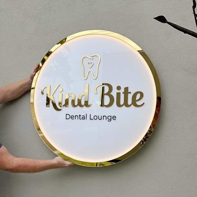 May include: A round sign with a gold trim and the words "Kind Bite Dental Lounge" in gold lettering. The sign has a white background with a gold tooth icon with a heart in the middle. The sign is being held by two hands.