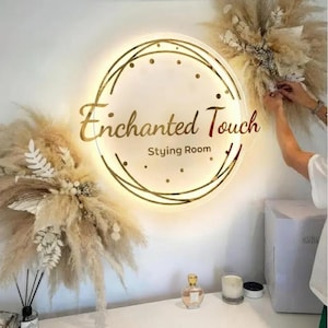 May include: A circular, illuminated sign with the text "Enchanted Touch Styling Room" in gold. The sign is framed by a gold geometric pattern and small lights. The image also shows dried floral arrangements and a perfume bottle.