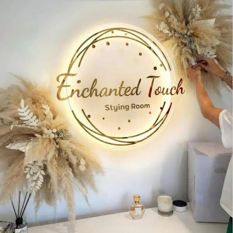 May include: A circular, illuminated sign with the text "Enchanted Touch Styling Room" in gold. The sign is framed by a gold geometric pattern and small lights. The image also shows dried floral arrangements and a perfume bottle.