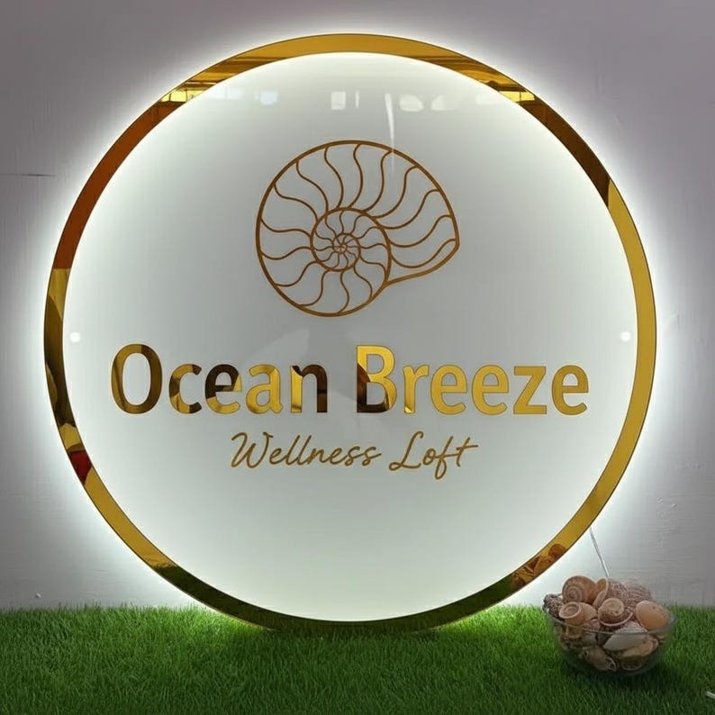 K&ouml;nnte beinhalten: Rundes Schild mit goldenem Rand und den Worten "Ocean Breeze Wellness Loft" in goldener Schrift. Ein goldenes Muscheldesign befindet sich &uuml;ber dem Text. Das Schild ist wei&szlig; mit einem leuchtenden Rand. Eine kleine Schale mit Muscheln befindet sich darunter.