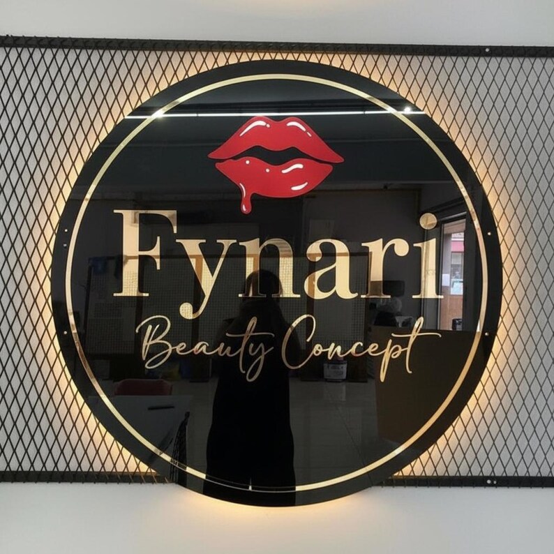 K&ouml;nnte beinhalten: Rundes schwarzes Schild mit goldenem Rand und den Worten "Fynari Beauty Concept" in goldener Schrift. Oben befindet sich eine rote Lippen-Grafik. Das Schild ist auf einem Metallgitter mit warmer Beleuchtung montiert.