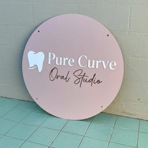 May include: A round, blush pink sign with the words "Pure Curve Oral Studio" in white and brown lettering. A white tooth graphic is on the left. The sign has four small holes and is leaning against a textured wall.