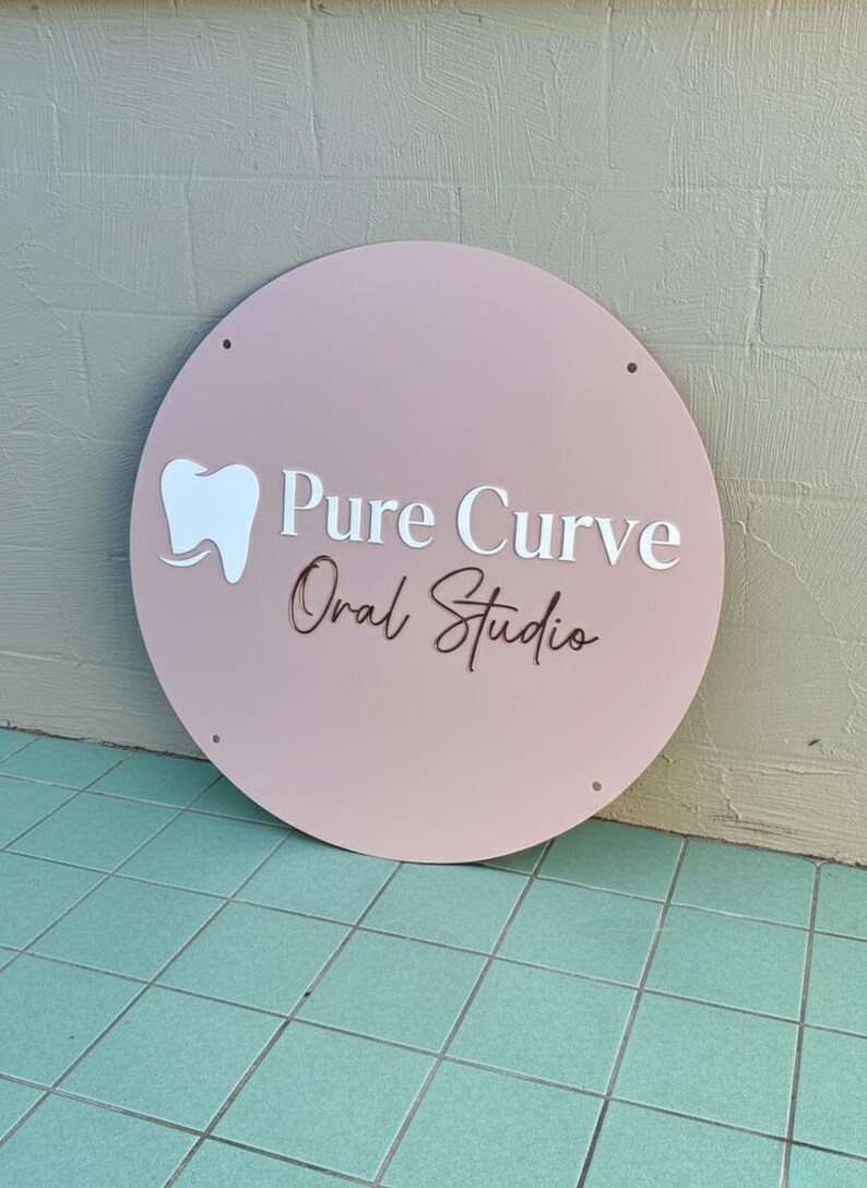 May include: A round, blush pink sign with the words "Pure Curve Oral Studio" in white and brown lettering. A white tooth graphic is on the left. The sign has four small holes and is leaning against a textured wall.