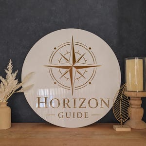 May include: A round, off-white wooden sign with a compass rose design in gold. The sign reads "HORIZON GUIDE" in gold lettering. The sign is displayed on a wooden surface with decorative items.