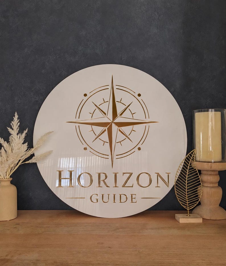 May include: A round, off-white wooden sign with a compass rose design in gold. The sign reads "HORIZON GUIDE" in gold lettering. The sign is displayed on a wooden surface with decorative items.