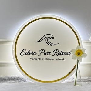 Puede incluir: Un letrero redondo e iluminado con el texto "Eclora Pure Retreat" y un logotipo de ola. El letrero tiene un borde dorado y un fondo blanco. Un narciso en un jarr&oacute;n de cristal est&aacute; en primer plano.
