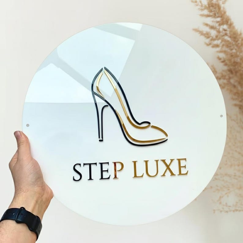 Pu&ograve; includere: Un cartello rotondo bianco con un logo di scarpa con tacco alto nero e oro. Il testo "STEP LUXE" &egrave; sotto il logo in nero e oro. Il cartello &egrave; tenuto da una mano.