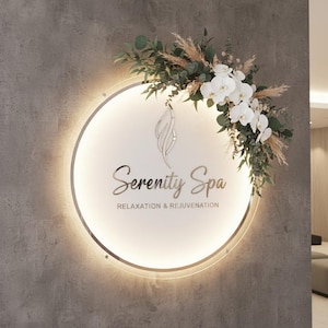 K&ouml;nnte beinhalten: Ein kreisf&ouml;rmiges, beleuchtetes Schild f&uuml;r "Serenity Spa" mit einem Blumengesteck. Das Schild zeigt den Namen des Spas und die Worte "Relaxation & Rejuvenation". Das Blumengesteck umfasst wei&szlig;e Orchideen, gr&uuml;nes Laub und getrocknete Gr&auml;ser.