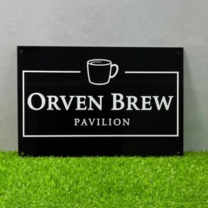 May include: A black rectangular sign with white text and a coffee cup graphic. The sign reads "Orven Brew Pavilion" in a bold, sans-serif font, with the cup above the text. The sign is mounted on a wall above green grass.