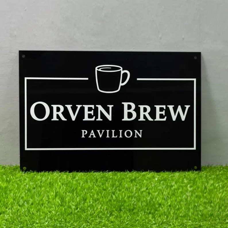 May include: A black rectangular sign with white text and a coffee cup graphic. The sign reads "Orven Brew Pavilion" in a bold, sans-serif font, with the cup above the text. The sign is mounted on a wall above green grass.