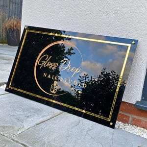 May include: A rectangular black sign with gold trim and the words "Gloss Drop Nail Studio" in gold script. The sign has a gold circle with a nail polish bottle icon. The sign is mounted on a stone surface.