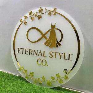 May include: A round sign with a white background and gold accents. The sign features the text "Eternal Style Co." in gold lettering, along with a gold dress silhouette and an infinity symbol. Gold ivy leaf accents adorn the edges.