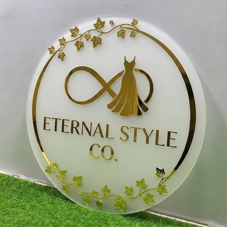 May include: A round sign with a white background and gold accents. The sign features the text "Eternal Style Co." in gold lettering, along with a gold dress silhouette and an infinity symbol. Gold ivy leaf accents adorn the edges.
