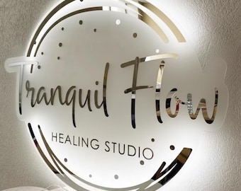 May include: A round, illuminated sign with the words "Tranquil Flow Healing Studio" in a mirrored, cursive font. The sign has a frosted glass appearance with a glowing white light and a mirrored border. Small dots are scattered around the text.