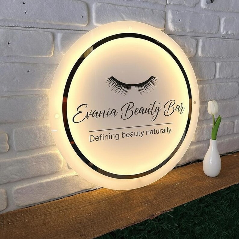 Puede incluir: Un letrero redondo e iluminado para Evania Beauty Bar, con un borde negro y el texto "Evania Beauty Bar" con el lema "Defining beauty naturally". El letrero incluye un gr&aacute;fico de pesta&ntilde;as. Un peque&ntilde;o jarr&oacute;n blanco con un tulip&aacute;n est&aacute; a la derecha.