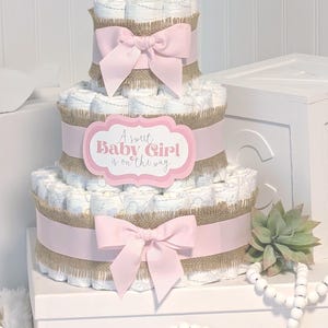 May include: A three-tiered diaper cake constructed from rolled white diapers with light blue patterns. Each tier is wrapped in a strip of brown burlap and a light pink ribbon with a bow. A pink sign reads A sweet Baby Girl is on the way.