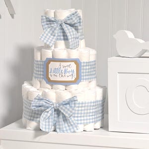 May include: A three-tiered diaper cake constructed from rolled white diapers, decorated with light blue and white gingham ribbons and bows. A rectangular sign on the middle tier features the text, A sweet Little Boy is on the way!